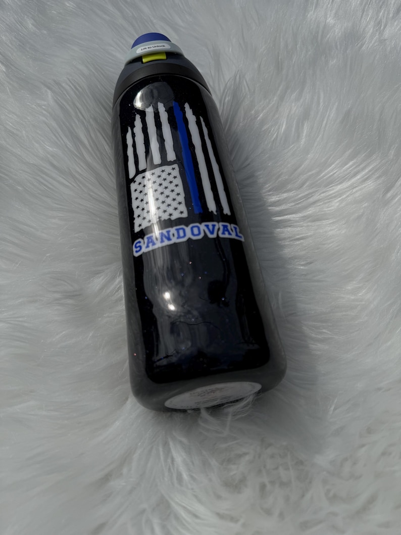 May include: Black water bottle with a blue and white American flag design and the name "Sandoval" in blue. The bottle has a blue lid and a black body.