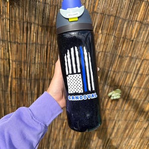 May include: Black water bottle with a blue and white graphic of the American flag and a blue line. The bottle has a blue lid and the name "SANDOVAL" is printed on the bottle.