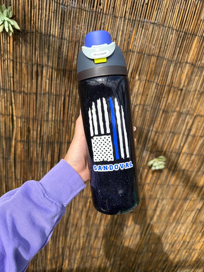 May include: Black water bottle with a blue and white graphic of the American flag and a blue line. The bottle has a blue lid and the name "SANDOVAL" is printed on the bottle.