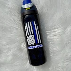 May include: A black water bottle with a blue and white flag design and the name "SANDOVAL" in white lettering. The bottle has a blue lid and a gray handle with the text "Lift to Unlock".