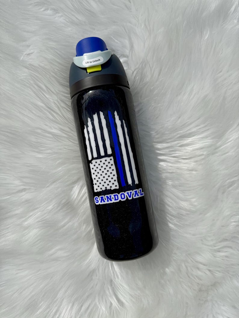 May include: A black water bottle with a blue and white flag design and the name "SANDOVAL" in white lettering. The bottle has a blue lid and a gray handle with the text "Lift to Unlock".