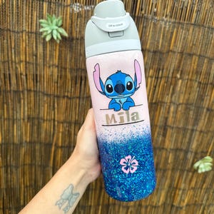 May include: A personalised water bottle with a cartoon Stitch design on a pink background. The lower half is blue glitter, with a pink flower. The name "Mila" is printed above the glitter. The bottle has a grey lid.