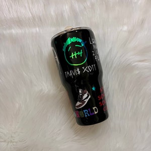 May include: Black stainless steel tumbler with a graphic design featuring a cartoon face, the text "Travis Scott", a trainer, and the text "Astroworld".