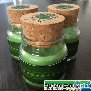 Puede incluir: Tres tarros de velas de vidrio verde con tapas de corcho. Cada tarro tiene una etiqueta verde y blanca con un patrón geométrico y el texto "SUPER Fam Mart" en japonés e inglés.