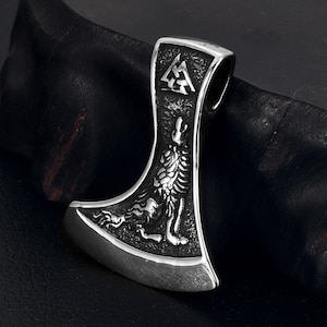 Puede incluir: Un colgante de hacha de plata con un diseño de lobo y un símbolo triangular en la parte superior. El colgante está sobre un fondo negro.