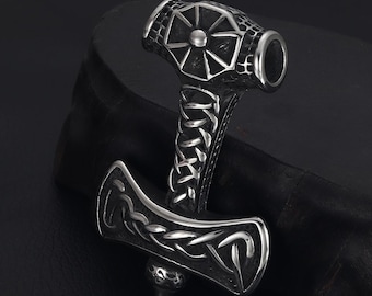 May include: A silver-coloured pendant in the shape of Thor's hammer, Mjolnir. The hammer features intricate Celtic knotwork and a detailed design on the head. The handle is wrapped in a chain-like pattern. The pendant is likely made of stainless steel.