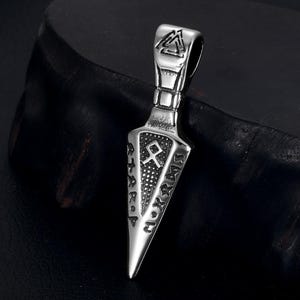 May include: A silver-coloured pendant in the shape of a dagger. The pendant features engraved runes and symbols, including a triangular design at the top. The dagger's blade is pointed, and the pendant has a loop for a chain.