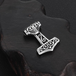 May include: A silver-coloured pendant shaped like Thor's hammer, Mjolnir. The hammer has detailed engravings on the head and handle, with a loop for a chain. The pendant is shown on a dark, textured surface.