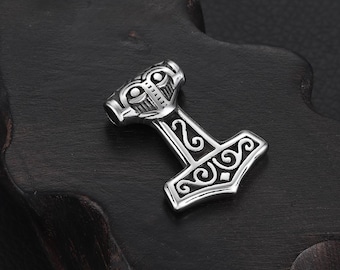 May include: A silver-coloured pendant shaped like Thor's hammer, Mjolnir. The hammer has detailed engravings on the head and handle, with a loop for a chain. The pendant is shown on a dark, textured surface.