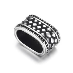 May include: A silver-coloured, oval-shaped bead with a textured, woven pattern. The bead has a central opening and is bordered by a dotted design. The bead is made of a shiny, metallic material.