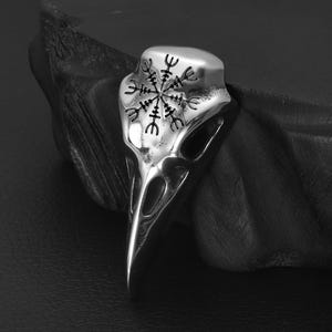 Peut inclure: Un pendentif en argent en forme de crâne d'oiseau avec un design détaillé. Le crâne a un motif circulaire au centre avec un symbole qui ressemble à une boussole.