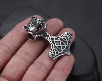 May include: A silver pendant shaped like Thor's hammer, Mjolnir. The hammer has a wolf head and Celtic knot detailing. The pendant is made of shiny metal, likely for a necklace. The design is intricate and detailed.