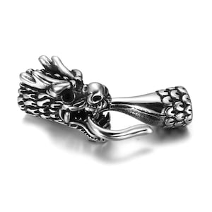 Może przedstawiać: Srebrny wisiorek w kształcie głowy smoka z misternymi detalami. Głowa smoka ma czaszkę, ostre zęby i teksturowane ciało. Wisiorek jest wykonany z błyszczącego metalu i prawdopodobnie jest elementem biżuterii.