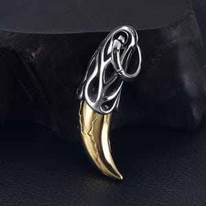 May include: A pendant featuring a gold-colored tooth design with a silver-colored, openwork top. The pendant has a ring for attaching to a chain. The background is dark.