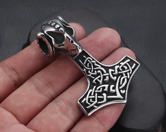 May include: A silver-coloured pendant shaped like Thor's hammer, featuring detailed knotwork and a ram's skull design at the top. The hammer is held in a hand against a dark backdrop. The pendant is a Viking-themed accessory.