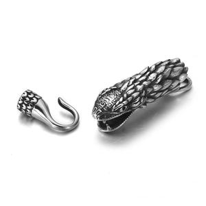 May include: A silver-coloured metal pendant featuring a detailed snake head with an open mouth and a separate hook-shaped clasp. The snake's head has textured scales and intricate facial details. The clasp has a textured pattern.