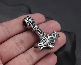 May include: A silver-coloured pendant shaped like Thor's hammer, with detailed engraved patterns. The hammer has a loop at the top for a chain. The design is intricate and the metal has a polished finish. A Viking-themed accessory.