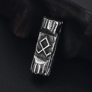 Peut inclure: Pendentif rectangulaire argenté à la texture vieillie. Il présente un symbole géométrique central stylisé et des lignes verticales en haut et en bas. Le design évoque un thème viking ou nordique.