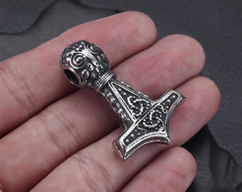 May include: A silver-coloured pendant shaped like Thor's hammer, Mjolnir. The pendant has detailed Celtic knotwork and decorative elements on the head and handle. The hammer is held in a hand against a dark background.