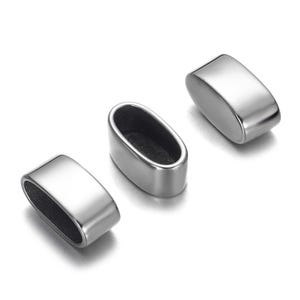 Pode incluir: Três contas de metal brilhantes, prateadas e ovais são exibidas sobre um fundo branco. Duas contas são sólidas, enquanto a terceira tem um centro aberto. As contas têm uma superfície lisa e reflexiva.