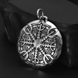 May include: A silver pendant with a detailed circular design featuring a central star and intricate swirling patterns. The pendant is on a black surface.