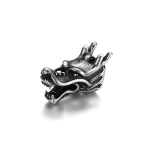 Pode incluir: Estatueta de cabeça de dragão prateada com escamas detalhadas, chifres e boca aberta. O dragão está voltado para a frente, com uma expressão feroz. A estatueta é feita de metal brilhante.