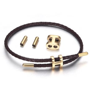 Pode incluir: Uma pulseira de couro castanho com detalhes em metal dourado. A pulseira apresenta uma faixa de couro trançado e um fecho dourado. Peças de metal dourado adicionais também estão incluídas.