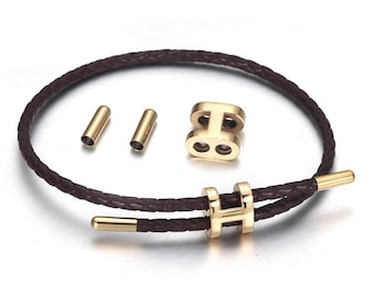 May include: A brown leather bracelet with gold-colored metal accents. The bracelet features a braided leather band and a gold-colored clasp. Additional gold-colored metal pieces are also included.