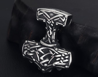 May include: A silver-coloured pendant shaped like Thor's hammer, Mjolnir. The hammer has detailed Celtic knotwork and geometric patterns on its head and handle. The pendant is shown on a dark surface.