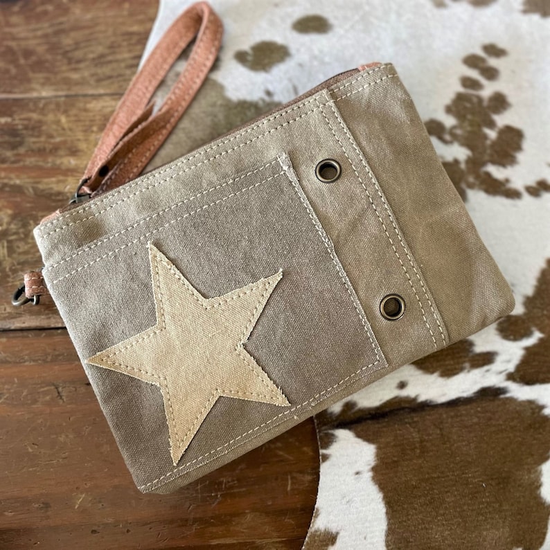 May include: A brown canvas zippered pouch with a large beige star applique. The pouch has a brown leather strap and two metal grommets.