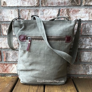 May include: A green canvas crossbody bag with a brown leather zipper pull and a brown leather flap. The bag has a zippered pocket on the front and a long adjustable strap. The bag is sitting on a wooden surface. RecycledMilitaryBags.com