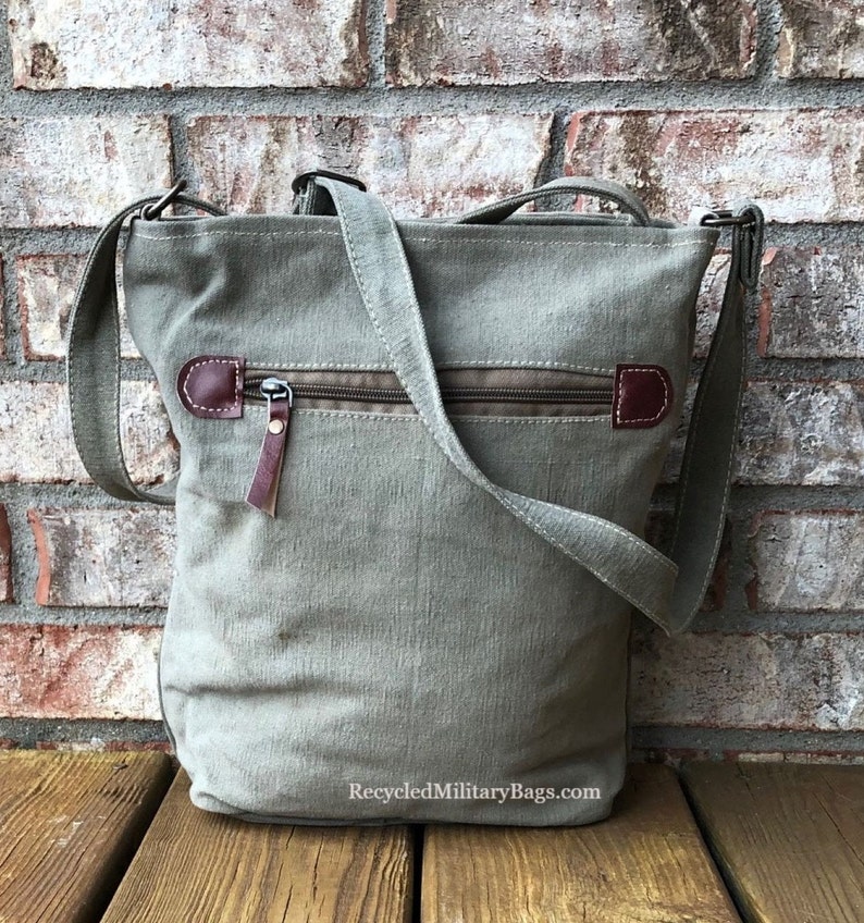 May include: A green canvas crossbody bag with a brown leather zipper pull and a brown leather flap. The bag has a zippered pocket on the front and a long adjustable strap. The bag is sitting on a wooden surface. RecycledMilitaryBags.com