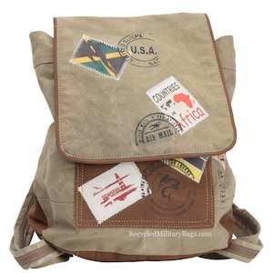 May include: A tan canvas backpack with brown leather accents and multiple patches. The patches feature images of airplanes, flags, and stamps with text like "U.S.A." and "Air Mail". The backpack has a flap closure and adjustable straps. RecycledMilitaryBags.com is printed on the bottom.