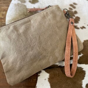 May include: A beige canvas clutch with a brown leather strap and zipper closure. The clutch is sitting on a white and brown cowhide surface.