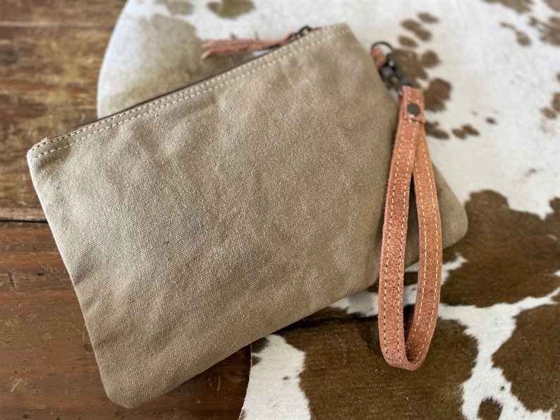 May include: A beige canvas clutch with a brown leather strap and zipper closure. The clutch is sitting on a white and brown cowhide surface.