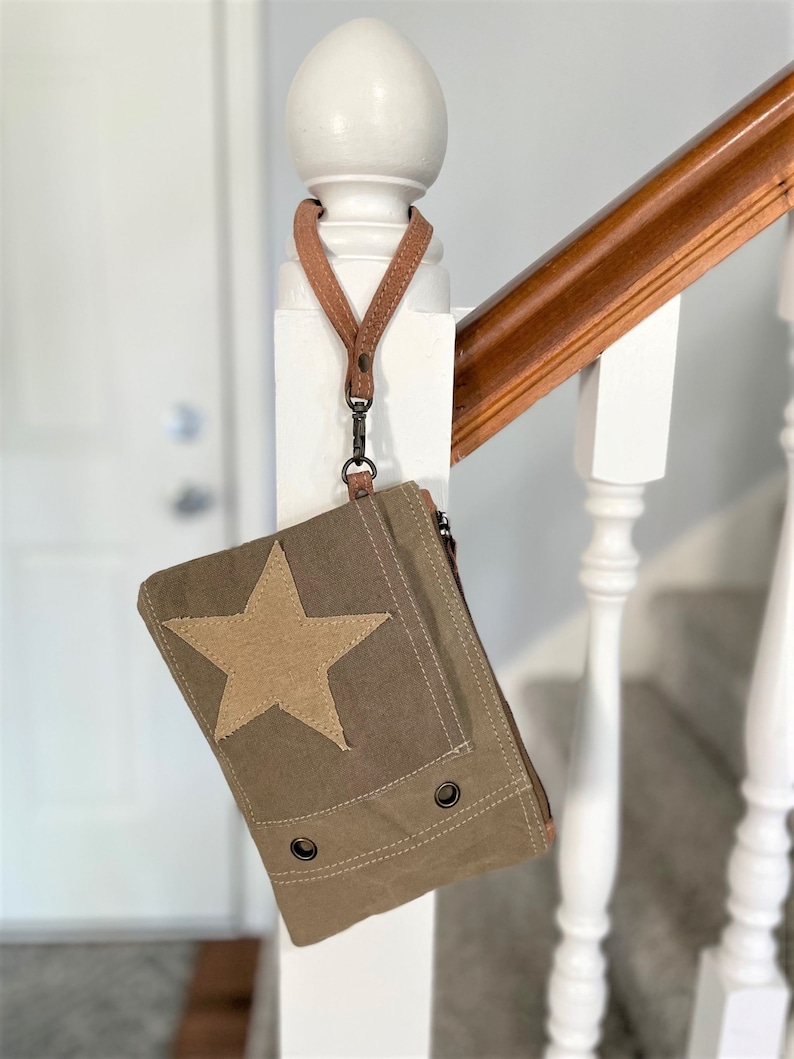 May include: A small, green canvas bag with a large tan star on the front. The bag has a brown leather strap and a zipper closure.