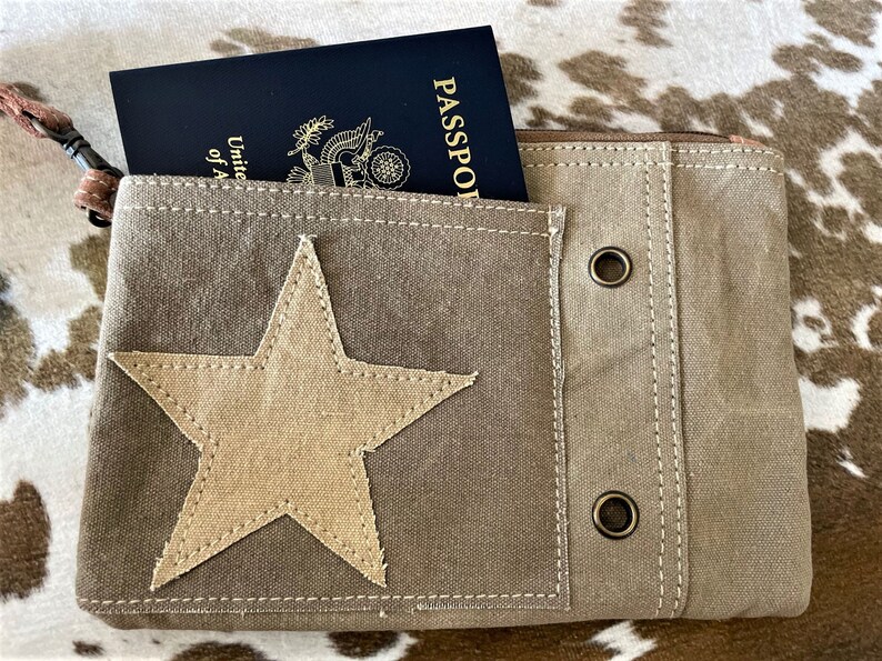 May include: A brown canvas travel pouch with a large beige star on the front. The pouch has a zipper closure and two metal eyelets on the side. A passport is visible inside the pouch.
