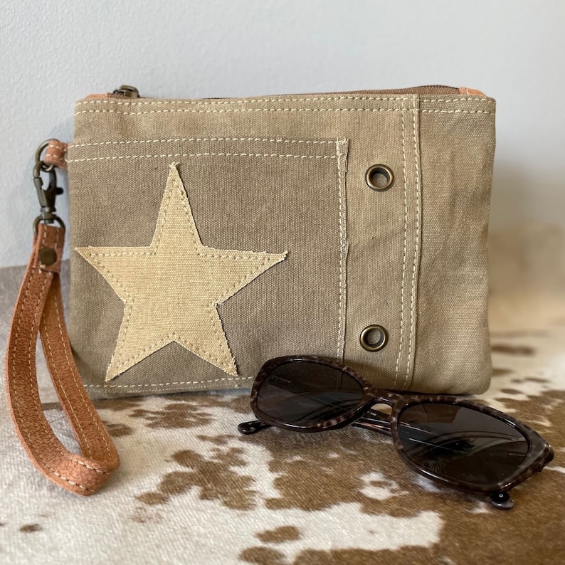 May include: A beige canvas clutch with a large white star and a brown leather strap. The clutch has two metal eyelets on the front. The clutch is sitting on a brown and white cowhide rug.