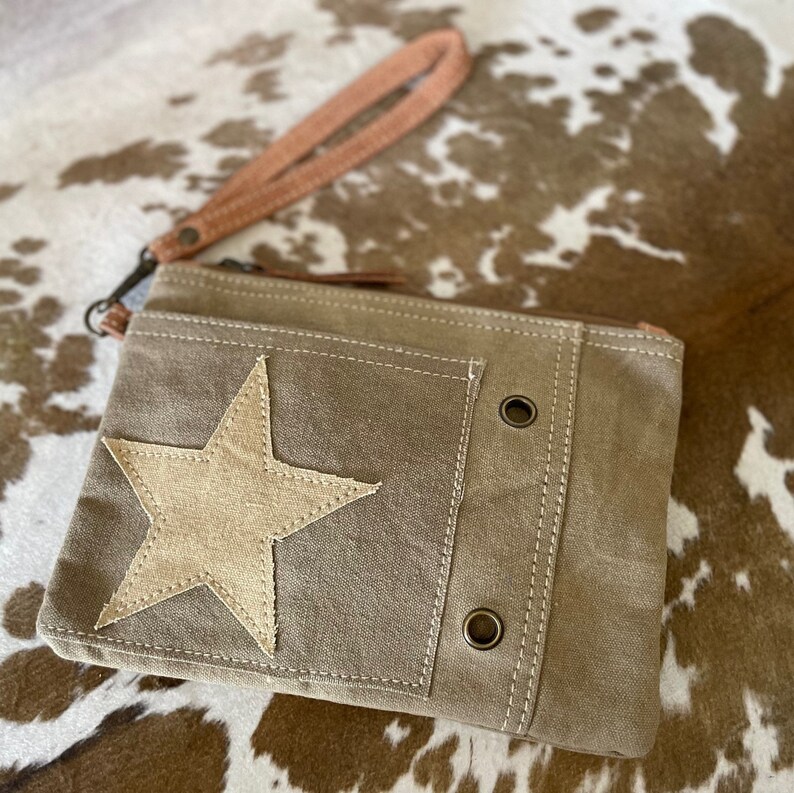 May include: A small, brown canvas clutch with a zipper closure and a star-shaped patch. The clutch has a brown leather strap and two metal eyelets on the front.