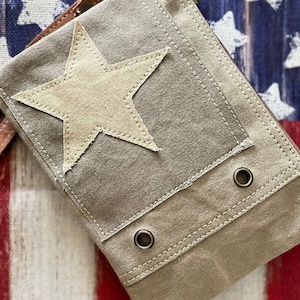 May include: A beige canvas bag with a large white star patch and two metal eyelets. The bag has a flap closure and a brown leather strap.