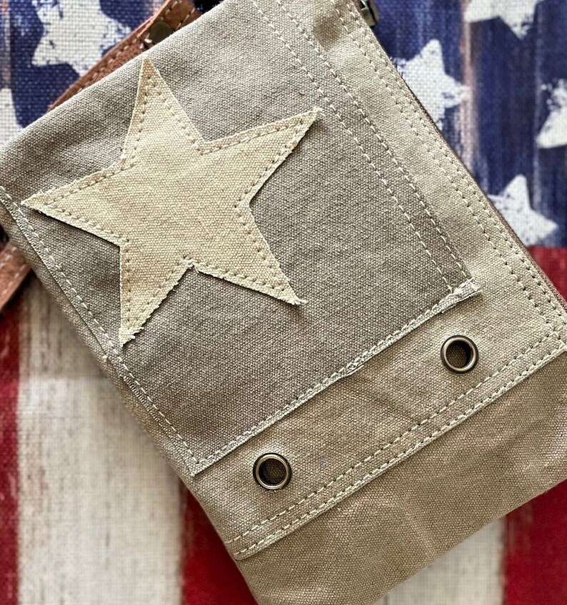 May include: A beige canvas bag with a large white star patch and two metal eyelets. The bag has a flap closure and a brown leather strap.