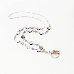 May include: A silver lanyard necklace with iridescent mother-of-pearl discs and small blue beads. The necklace has a chain and a clasp for attaching keys or an ID badge. The design is elegant and suitable for everyday wear.