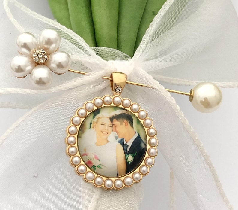 May include: A gold-toned circular photo pendant featuring a couple in wedding attire, framed by small white pearls. The pendant is attached to a gold-toned lapel pin adorned with a white organza ribbon bow and a five-petaled faux pearl flower.