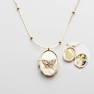 May include: A gold chain necklace with a white oval locket featuring a butterfly design. The locket opens to reveal a space for a photo.
