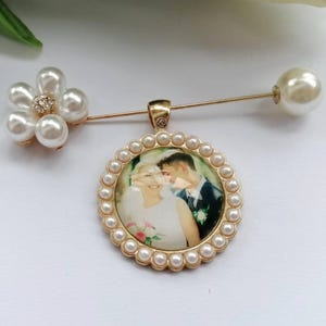 May include: A gold-toned brooch pin featuring a circular pendant with a photograph of a couple, surrounded by a border of small white pearls. A flower-shaped accent made of five white pearls and a central crystal sits on the pin stem.