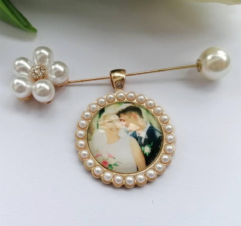 May include: A gold-toned brooch pin featuring a circular pendant with a photograph of a couple, surrounded by a border of small white pearls. A flower-shaped accent made of five white pearls and a central crystal sits on the pin stem.