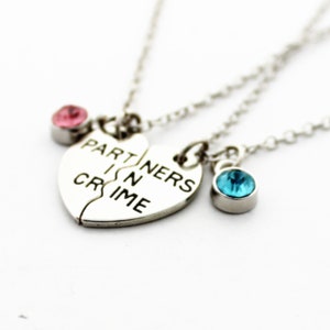 May include: Silver-toned necklace set featuring a split heart pendant with the words "PARTNERS IN CRIME". The set includes two additional necklaces, each with a small gemstone, one pink and one blue, on a delicate chain.