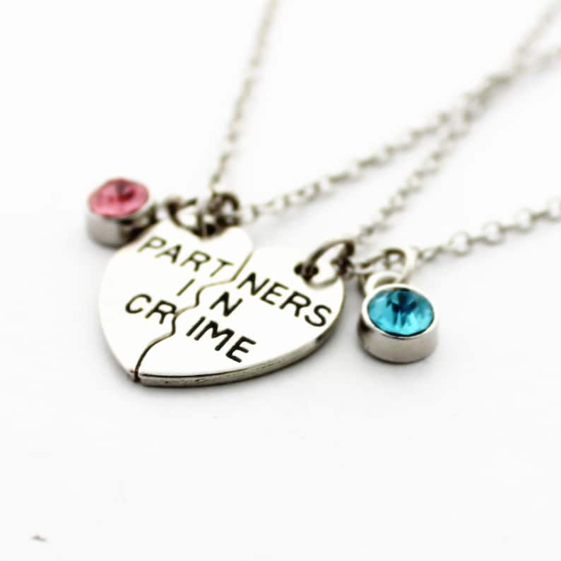May include: Silver-toned necklace set featuring a split heart pendant with the words "PARTNERS IN CRIME". The set includes two additional necklaces, each with a small gemstone, one pink and one blue, on a delicate chain.