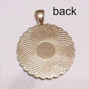 May include: A round, gold-toned metal pendant featuring a scalloped edge and a textured, radiating geometric pattern across its surface. The center is a smooth, circular flat area. A small bail is attached to the top. The text back is visible.