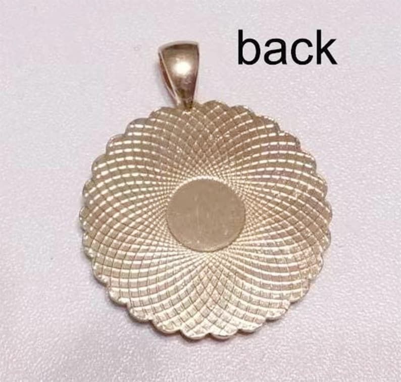 May include: A round, gold-toned metal pendant featuring a scalloped edge and a textured, radiating geometric pattern across its surface. The center is a smooth, circular flat area. A small bail is attached to the top. The text back is visible.